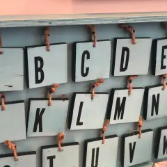 Drewniane tabliczki z literami alfabetu angielskiego pisanego, wiszące na sznurkach. Widoczne litery A, B, C, D, E, J, K, L, M, N, O, U, V, W.