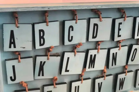Drewniane tabliczki z literami alfabetu angielskiego pisanego, wiszące na sznurkach. Widoczne litery A, B, C, D, E, J, K, L, M, N, O, U, V, W.