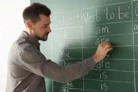 Nauczyciel pisze na tablicy odmianę czasownika "to be". Nauka angielskiego na poziomie C1 to cel wielu uczniów.