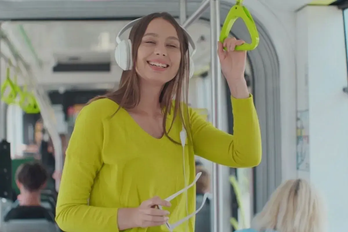 Uśmiechnięta dziewczyna w słuchawkach słucha podcastów do nauki angielskiego w tramwaju, trzymając się poręczy.